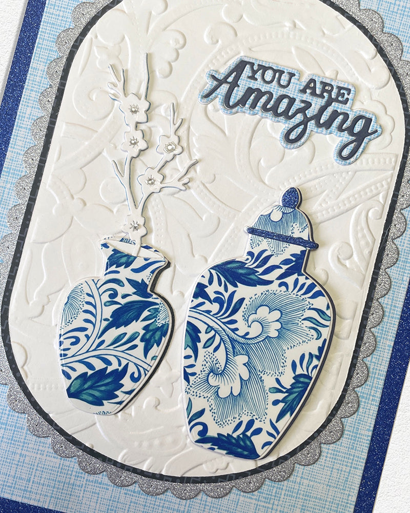 Close-up detail of a handmade luxury card showing the upper portion of a scalloped ivory oval frame with hand-embossed texture, two blue and white chinoiserie ginger jar illustrations, a delicate branch spray, and the sentiment "YOU ARE Amazing" in layered bold and script lettering. Handmade greeting card made in New Zealand.