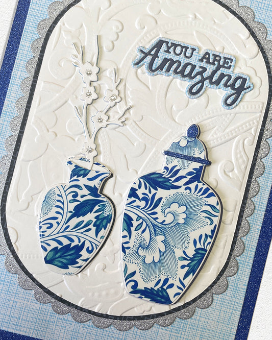 Close-up detail of a handmade luxury card showing the upper portion of a scalloped ivory oval frame with hand-embossed texture, two blue and white chinoiserie ginger jar illustrations, a delicate branch spray, and the sentiment "YOU ARE Amazing" in layered bold and script lettering. Handmade greeting card made in New Zealand.