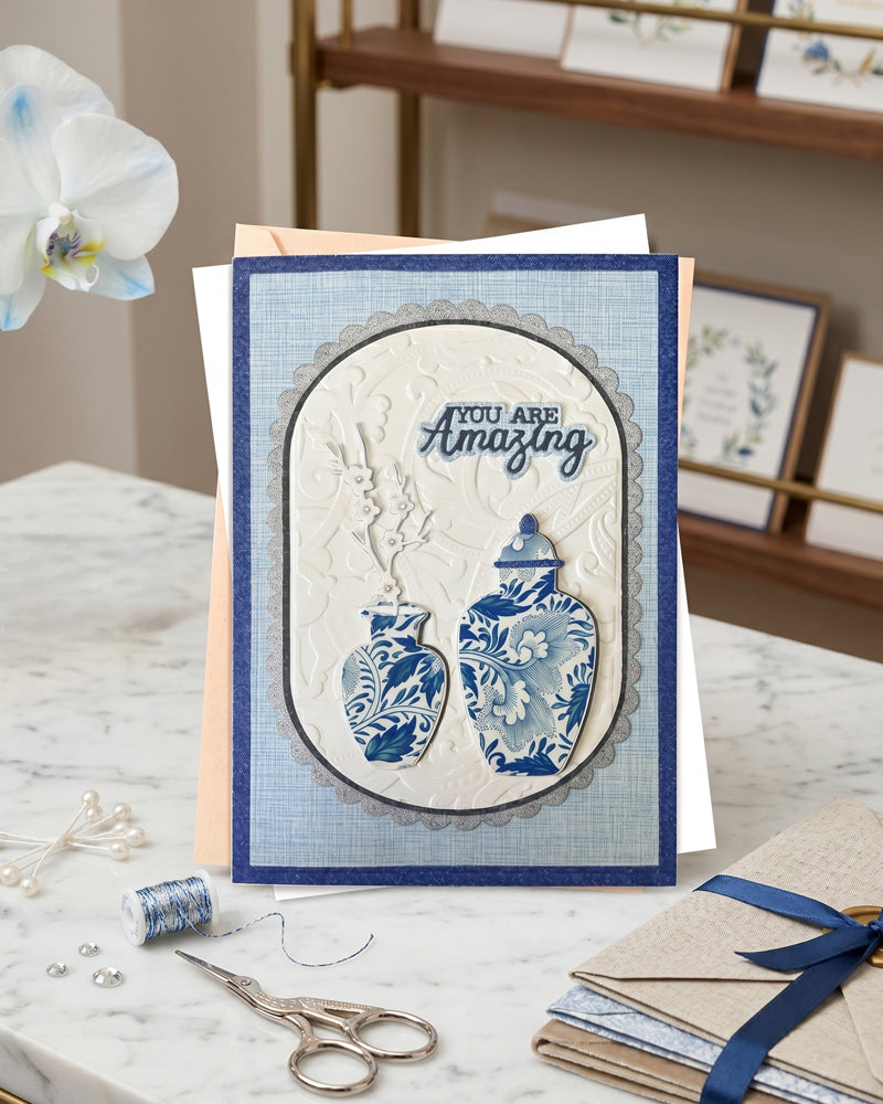 Handmade navy and ivory chinoiserie-style greeting card displayed upright in a bright styled setting with scissors and soft props alongside. Classic blue and white tones with elegant embossed detailing. A sophisticated handmade card made in New Zealand by Del Cards.