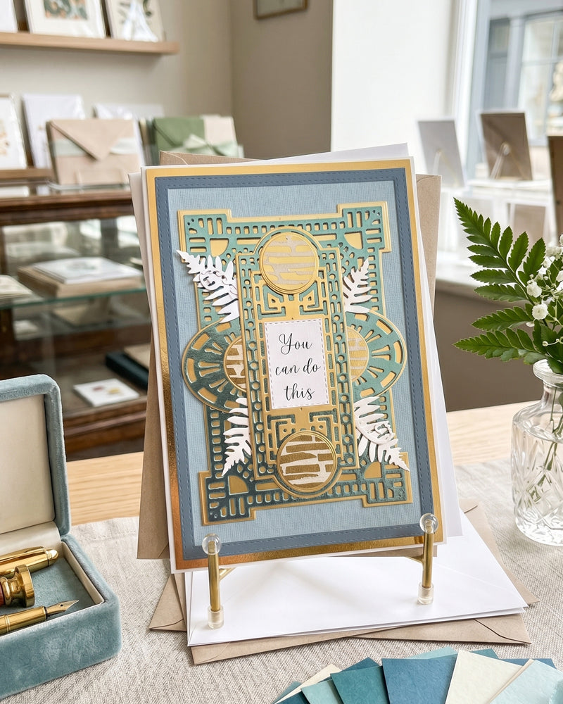 Handmade gold and blue "You can do this" encouragement card displayed upright on a wooden easel in a bright styled interior setting with a fern plant alongside. Steel blue, sage and gold tones with silver fern detail make this a striking handmade card made in New Zealand by Del Cards NZ.