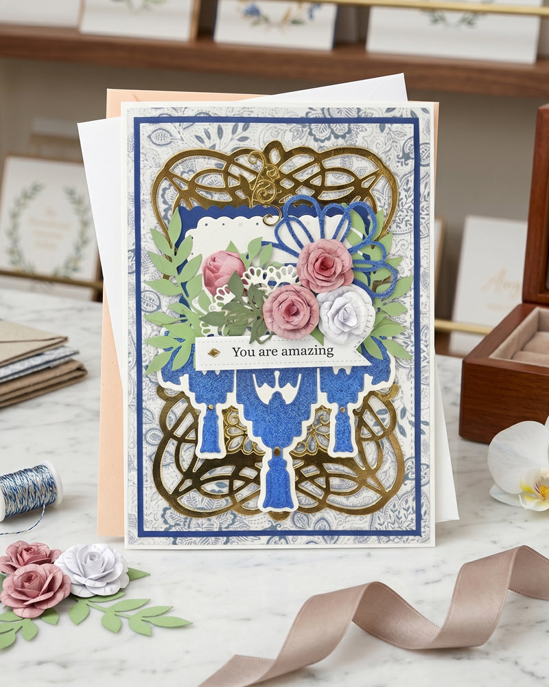 Handmade luxury greeting card with an intricate gold and black swirling filigree die-cut frame layered over a white embossed background. A bold cobalt blue glitter cardstock panel runs vertically through the centre, topped with an ornate blue chandelier-style die-cut. Lush hand-applied paper roses in pink, blush and white with green leaves cascade from the upper centre. Sentiment reads "You are amazing" in elegant script on a small tag. Handmade luxury card made in NZ by Del Cards NZ